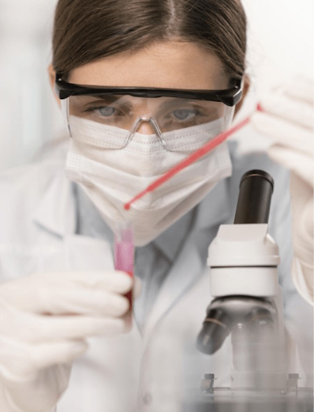 Una científica está dejando caer gotas de un elemento en un tubo de ensayo, lleva protección para los ojos y una mascarilla