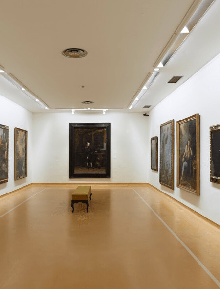 Una sala de un museo, podemos ver cuadros en las tres paredes y un banco en el centro de la sala