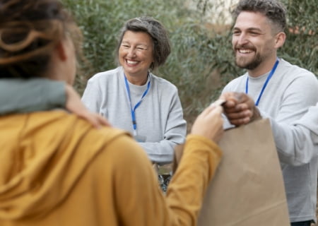 Dos cooperantes, con sudaderas grises y un identificador con cordón azul, ayudan a repartir comida en bolsas de papel