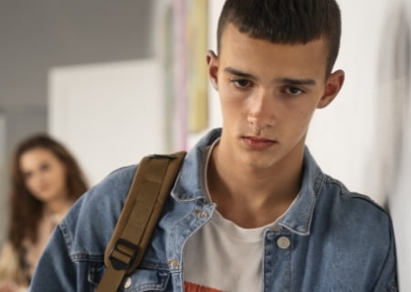 Un chico joven de cara triste, con una camiseta blanca, una chaqueta tejana y una mochila se apoya en una pared