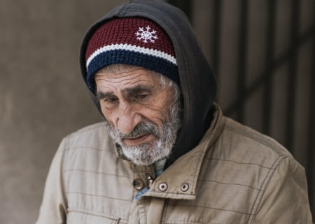 Un anciano con cara triste, tiene una barba canosa, la piel ajada y viste ropas viejas para protegerse del frío