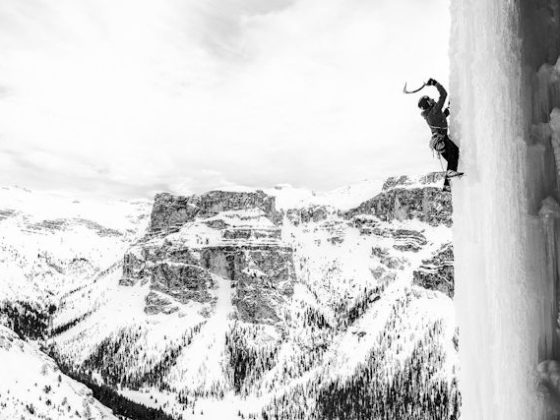 En un primer plano, sobre una pared de hielo vemos a un escalador usando un piolet para poder subir. Al fondo un macizo montañoso cubierto de nieve