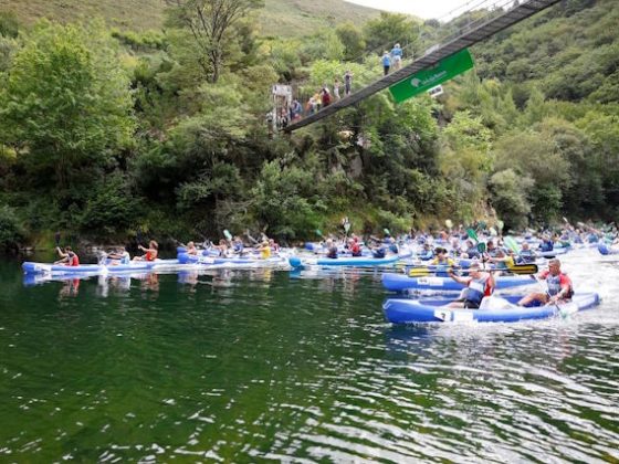Varias piraguas pasan bajo un puente que cruza el Río Sella, cuya orilla está cubierta por árboles, sobre el puente vemos público