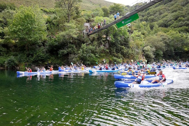 Varias piraguas pasan bajo un puente que cruza el Río Sella, cuya orilla está cubierta por árboles, sobre el puente vemos público